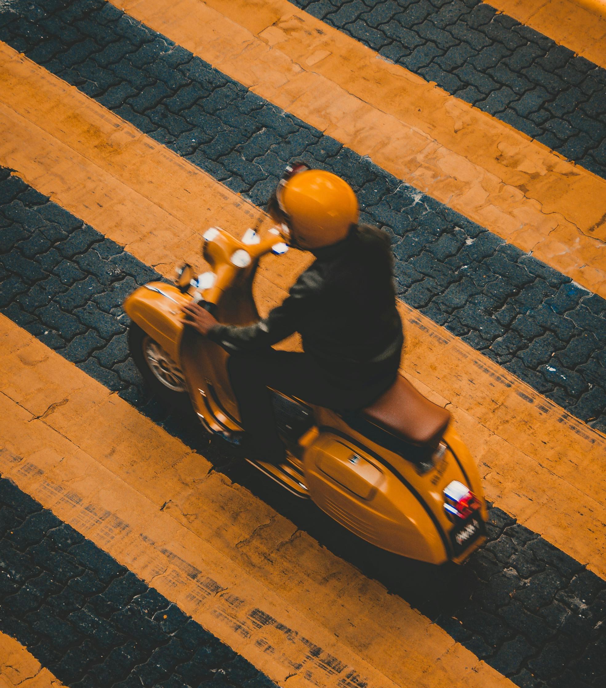 Person fährt auf einer gelben Vespa zwischen den Streifen eines gelben Fußgängerübergangs hindurch. Sie trägt außerdem einen gelben Helm. 