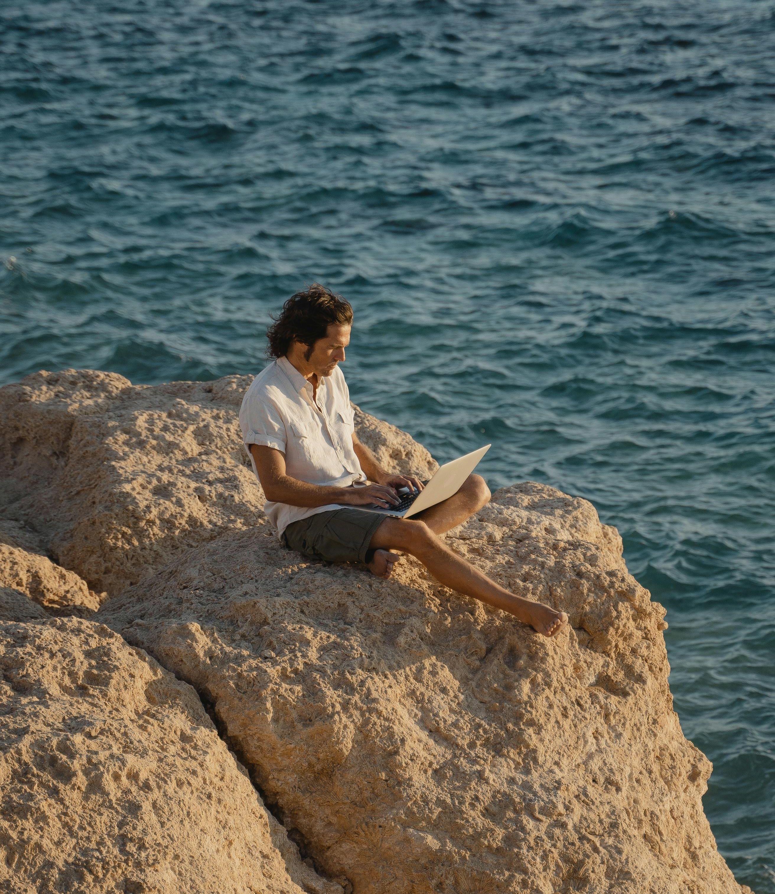 Ein Mann sitzt auf einem Felsen am Meer und arbeitet von seinem Laptop aus. 